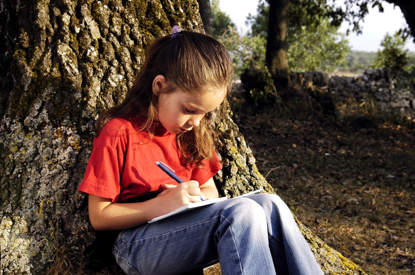 young girl is sitting under a tree and is writing in her notebook